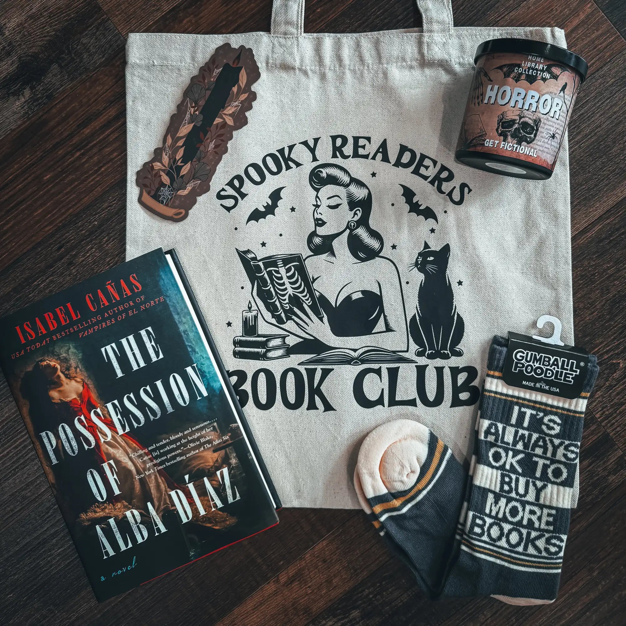 Beige canvas tote bag with black ink print of a pin-up woman reading beside a cat, surrounded by bats and the text ’spooky readers book club’