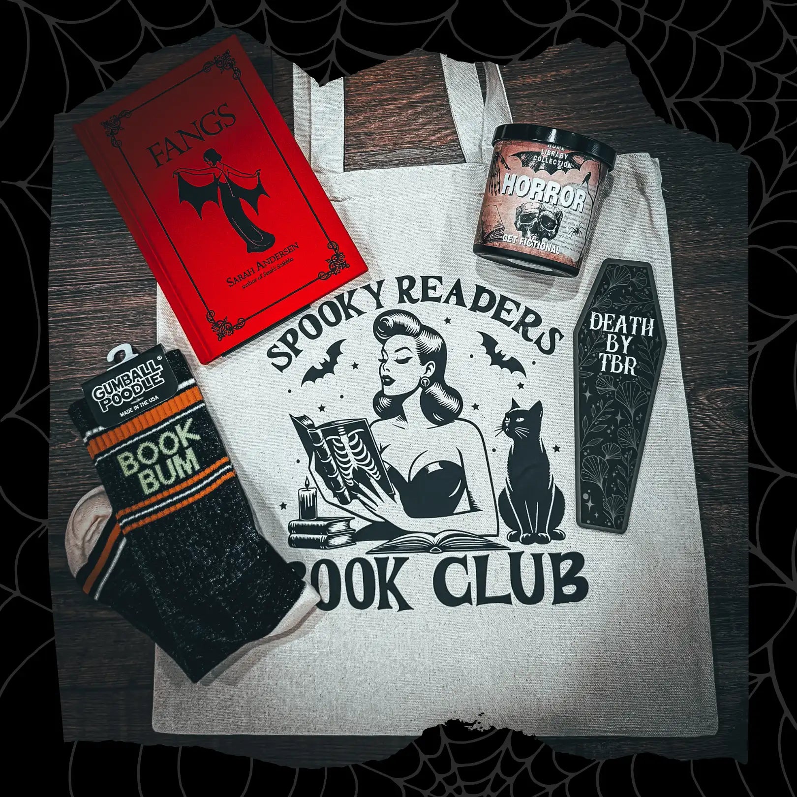 A beige canvas tote bag featuring a black ink illustration of a vintage-style woman reading with bats, stars, and a cat beside her.